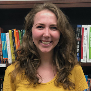 Jenna Kahn is a white woman with shoulder-length brown hair and brown eyes. She is standing in front of a wooden bookshelf full of books. She is wearing a gold-colored short-sleeved shirt.